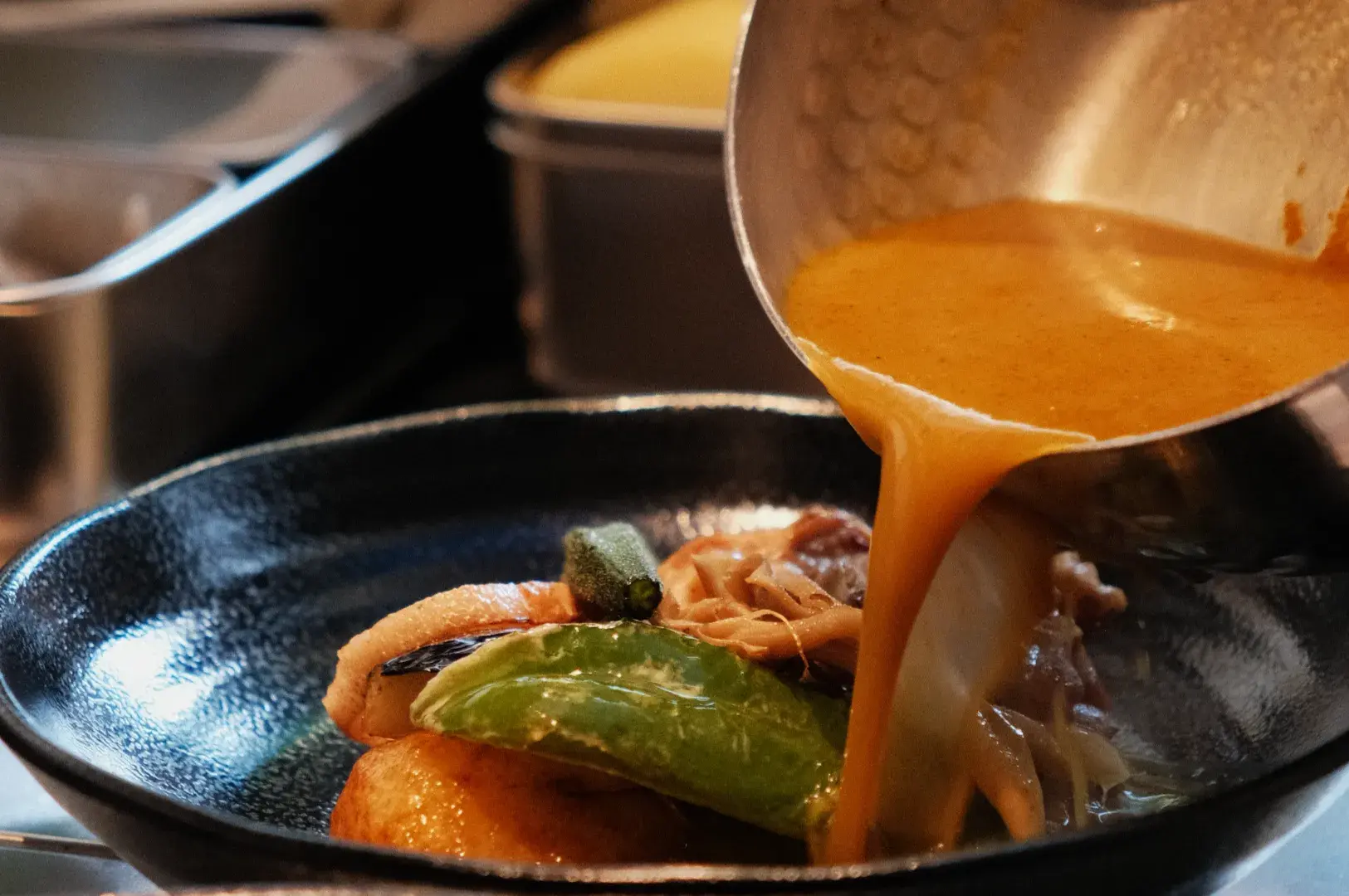 A chef pouring a rich sauce over a plate of food, featuring vegetables and meat, in a kitchen setting.