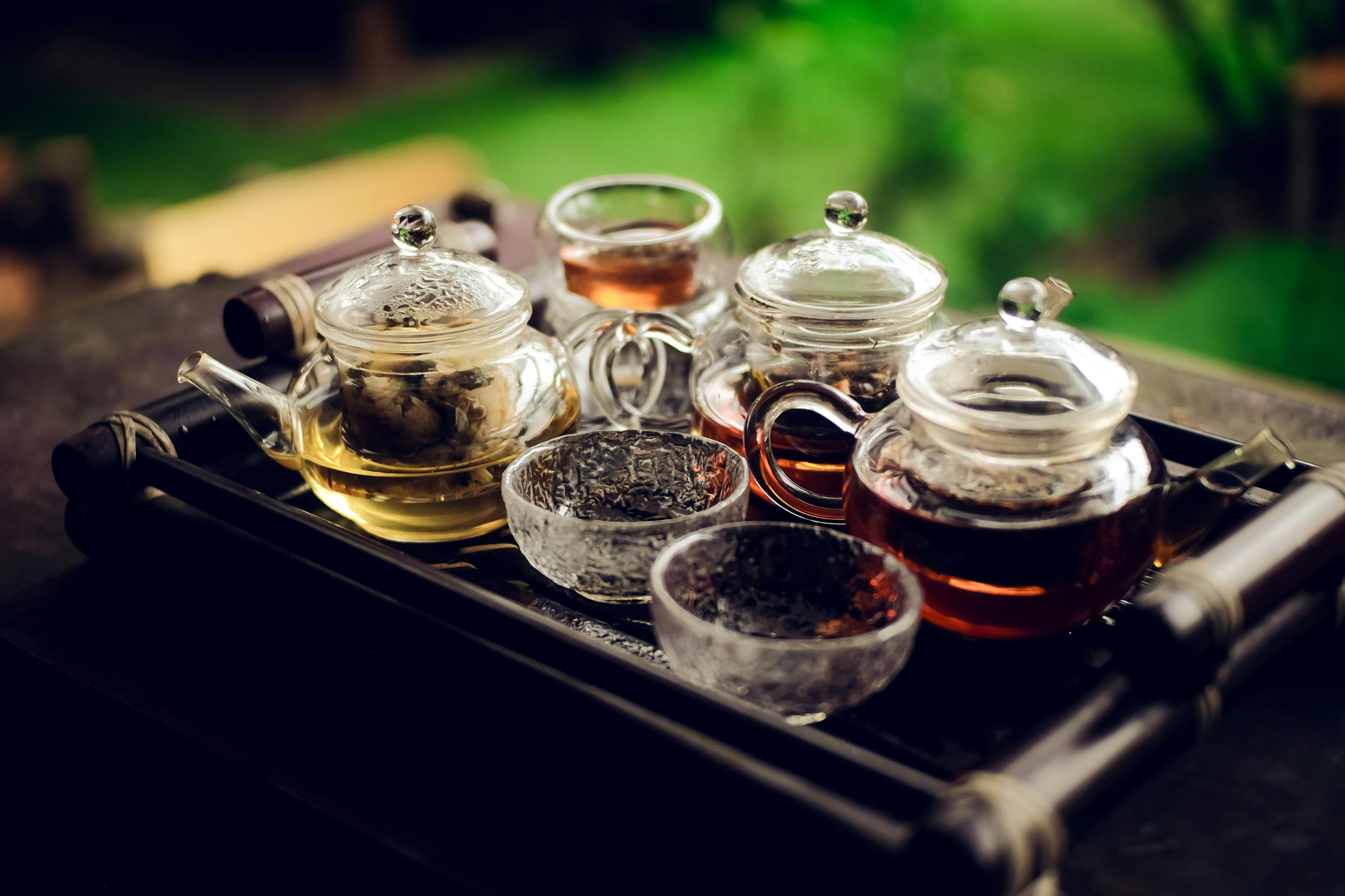 Three glass teapots filled with varying shades of tea are arranged on a dark, bamboo-style tray. Several small, matching glass cups accompany the pots, all set against a lush, blurred green background.