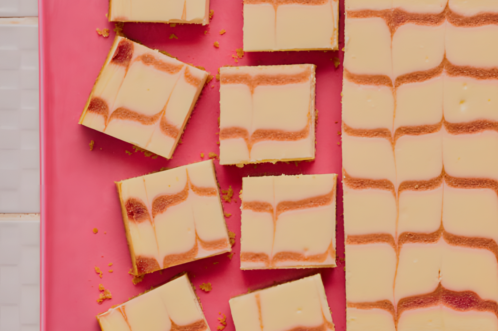 Close-up of cheesecake bars on a pink surface, each with a smooth cream layer and wavy caramel lines. The bars are neatly arranged, evoking a playful, sweet ambiance.