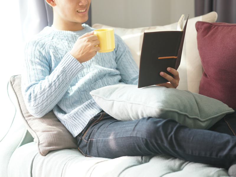 Homem toma café ou café com leite enquanto lê um livro em casa. Imagem: PP para Canva Pro.
