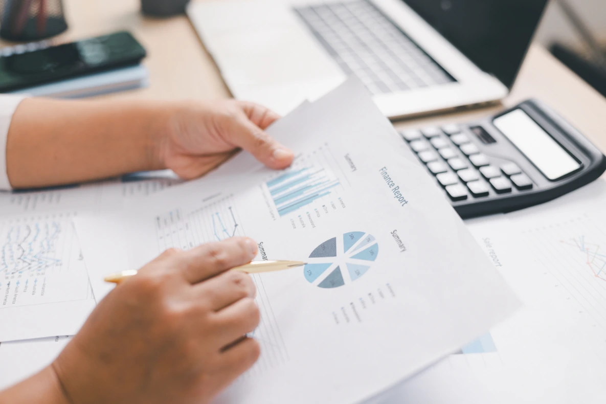 A person is reviewing financial reports with charts and a pie graph, with a calculator and laptop in the background.