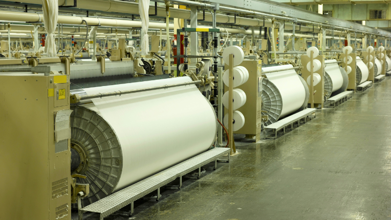 A long row of industrial looms with large white rolls of fabric and spools of thread in a factory setting.