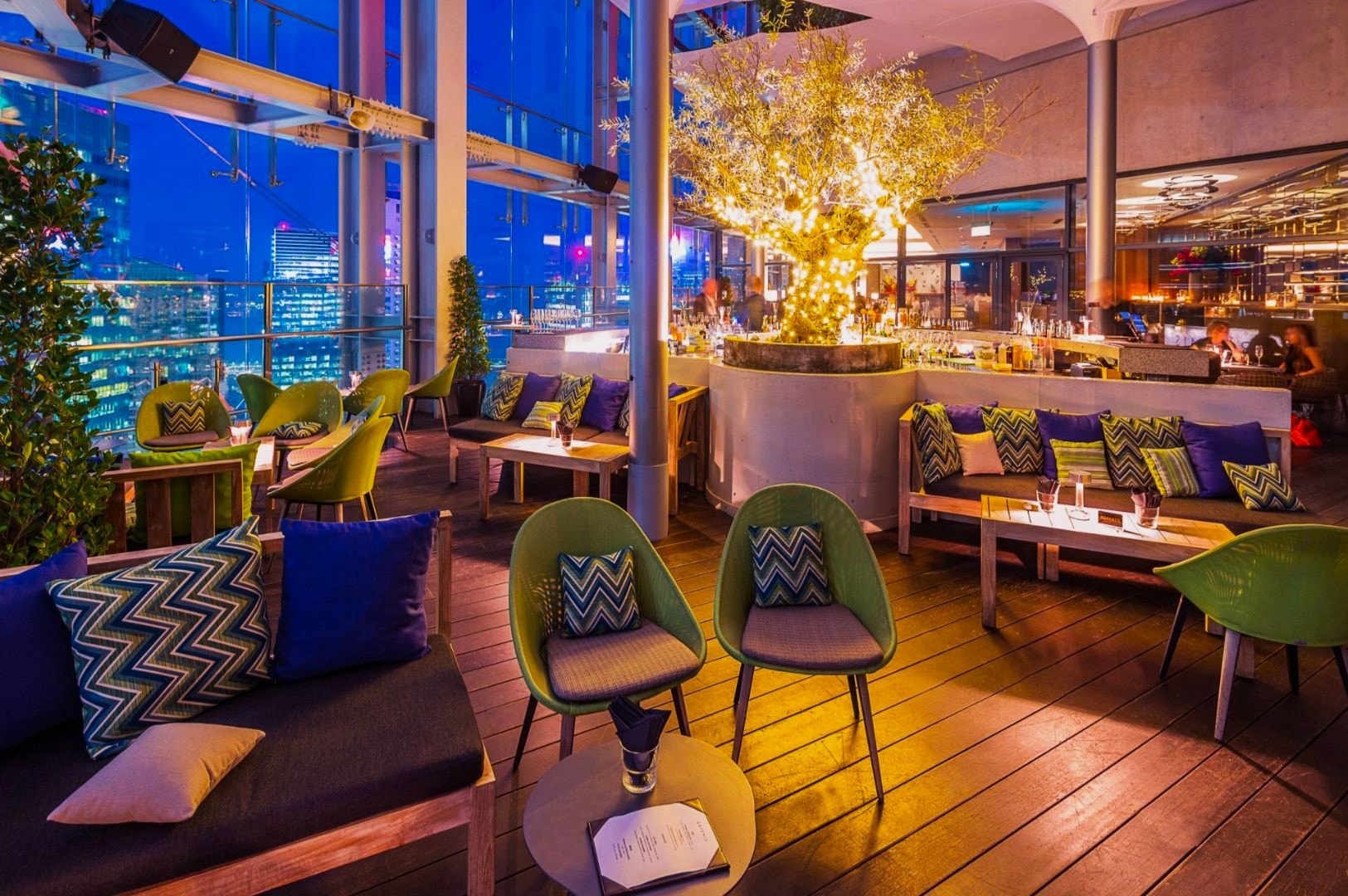 Modern rooftop lounge at night with city views. Ambient lighting, cozy seating with colorful cushions, and a lit tree create a relaxed, elegant atmosphere.