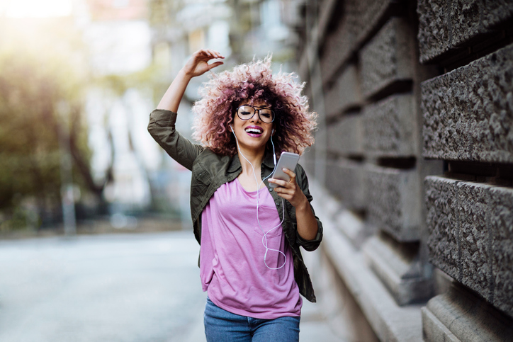 Super happy young woman with curly hair and glasses smiling as she walks down the street holding her phone .