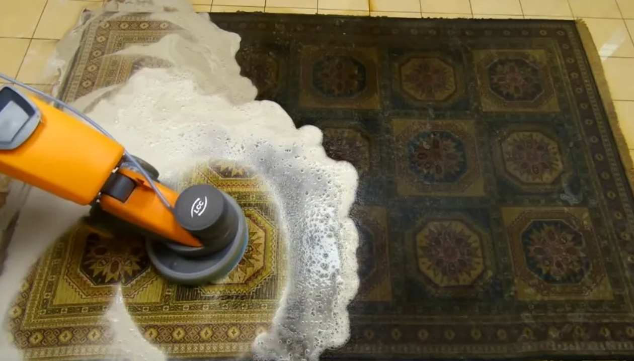 An orange rotary floor cleaning machine scrubbing a heavily soiled patterned rug, with visible soap foam showing a stark contrast between the cleaned and dirty sections of the rug. The cleaning process reveals vibrant gold and brown geometric designs beneath the grime