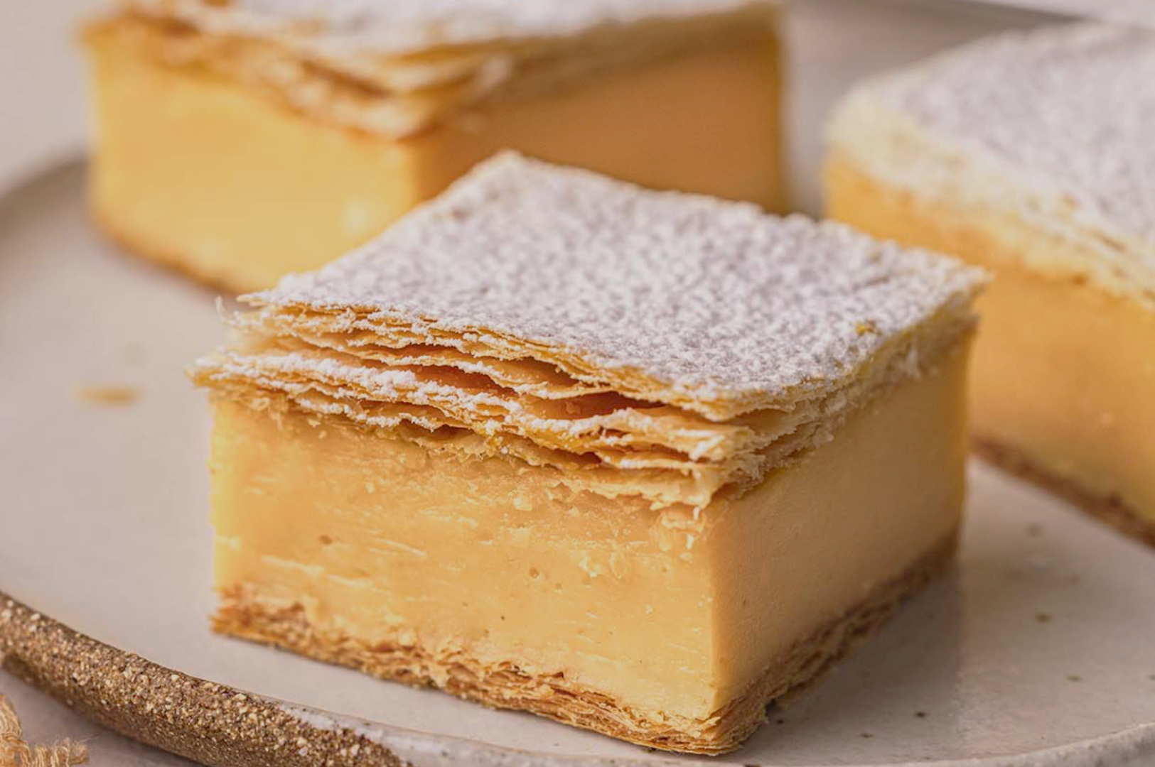 A creamy vanilla slice with flaky puff pastry layers, topped with powdered sugar. The dessert sits on a white plate, exuding a decadent, indulgent feel.