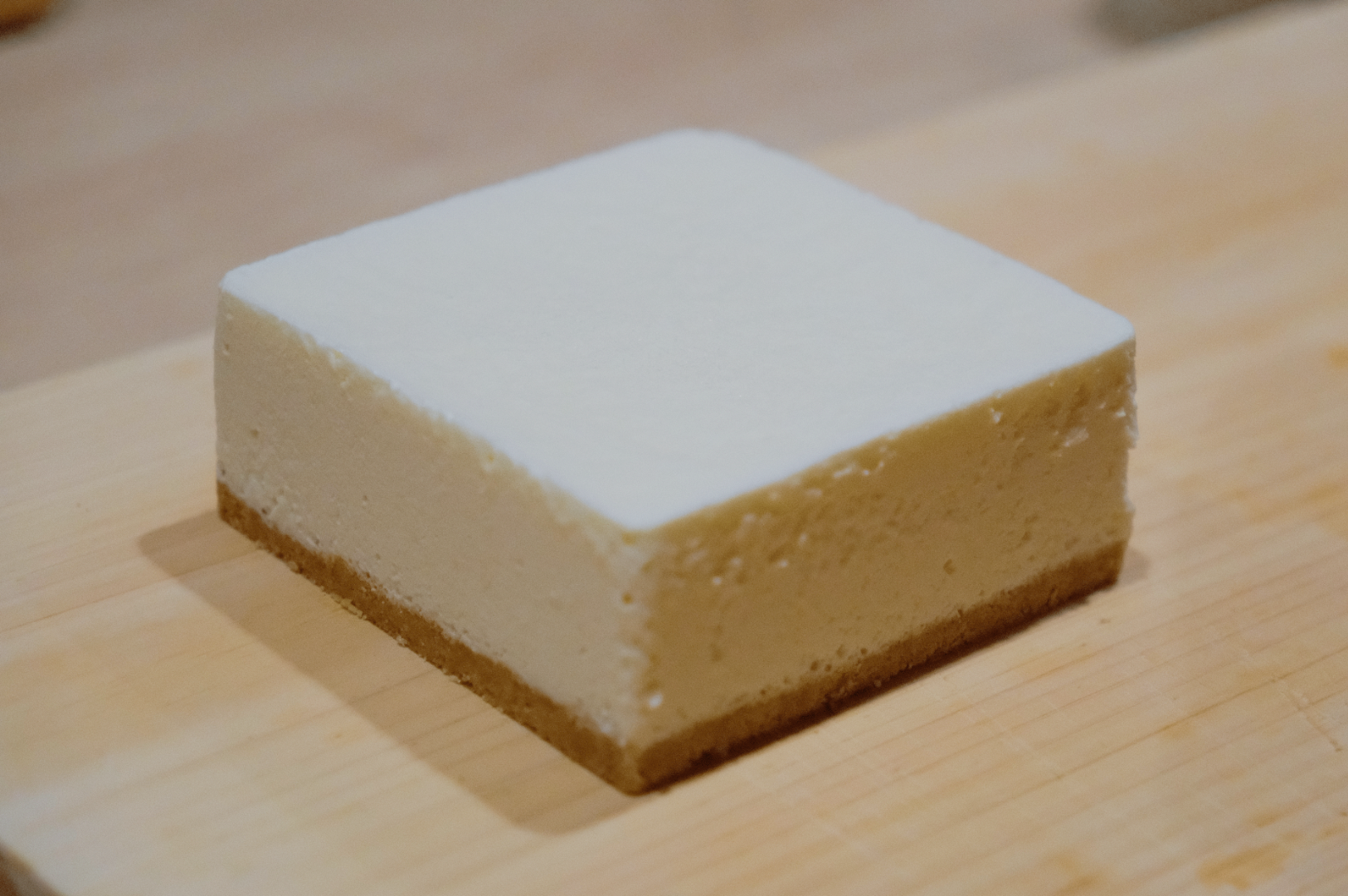 A square slice of creamy cheesecake with a smooth white top and a light brown crust sits on a wooden surface. The image conveys simplicity and deliciousness.