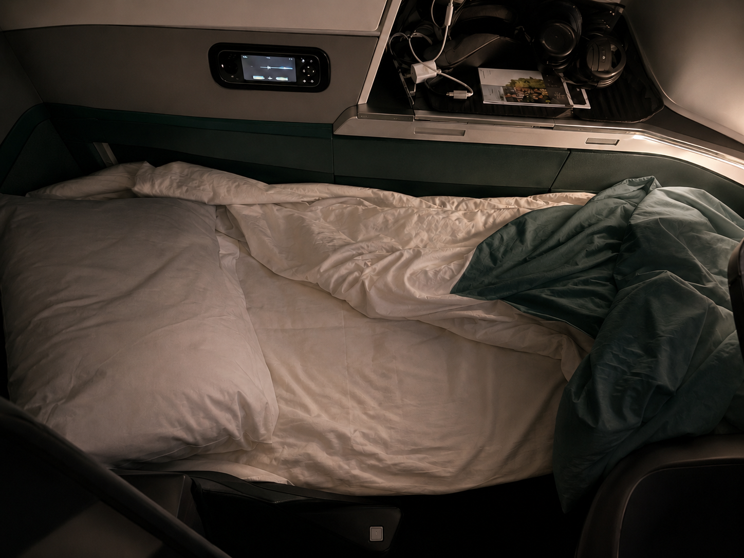 Business class lie-flat bed with white bedding and deep green accents, inspired by Cathay Pacific’s cabin colors, with headphones and storage beside the seat.