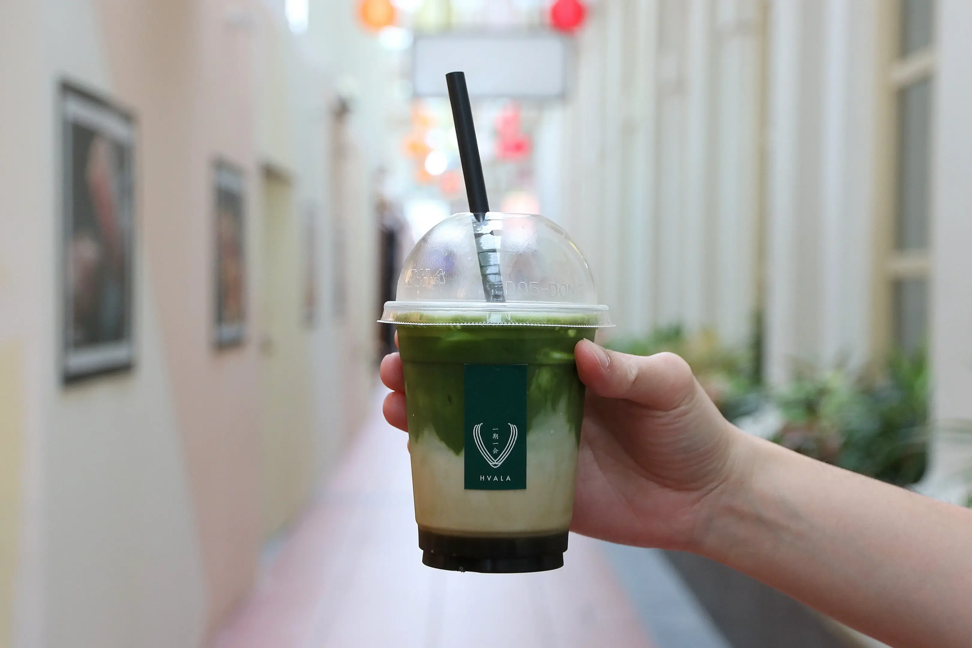 A person's hand holds a layered iced matcha latte from Hvala in a clear plastic cup with a black straw. The drink is positioned in the foreground against a softly blurred, narrow outdoor walkway lined with framed art and colorful decorations.