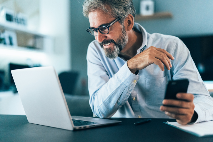 Gray-haired man with dark glasses smiling and working on his laptop.