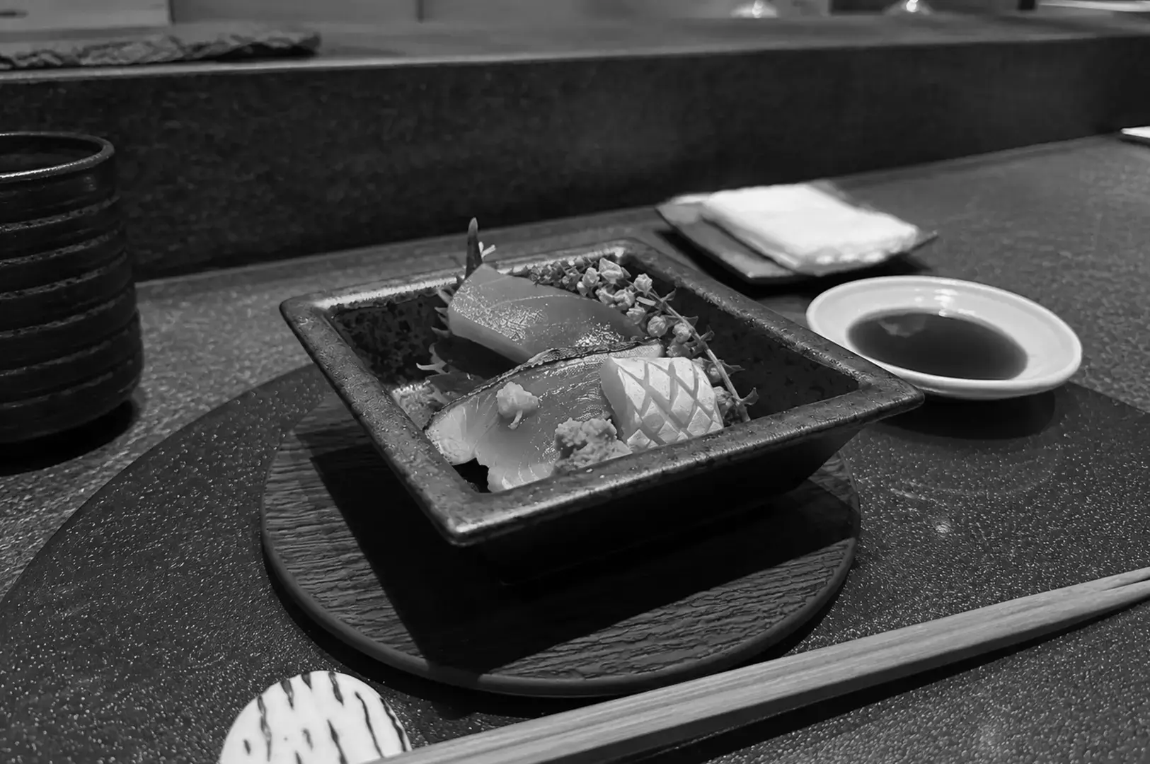 Sashimi pieces served in a square ceramic dish on a dark plate beside soy sauce, chopsticks, and stacked bowls.