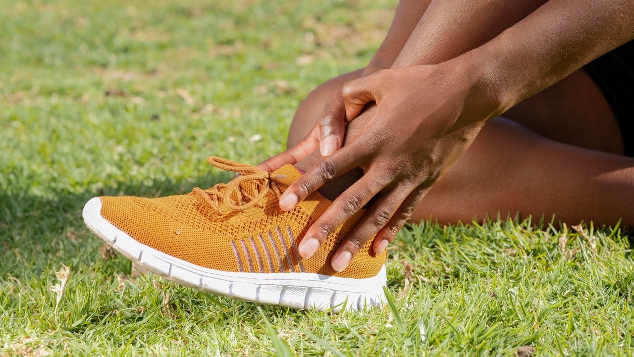 Runner holding sore foot while wearing sneakers on grass, showing shoe related foot pain after activity