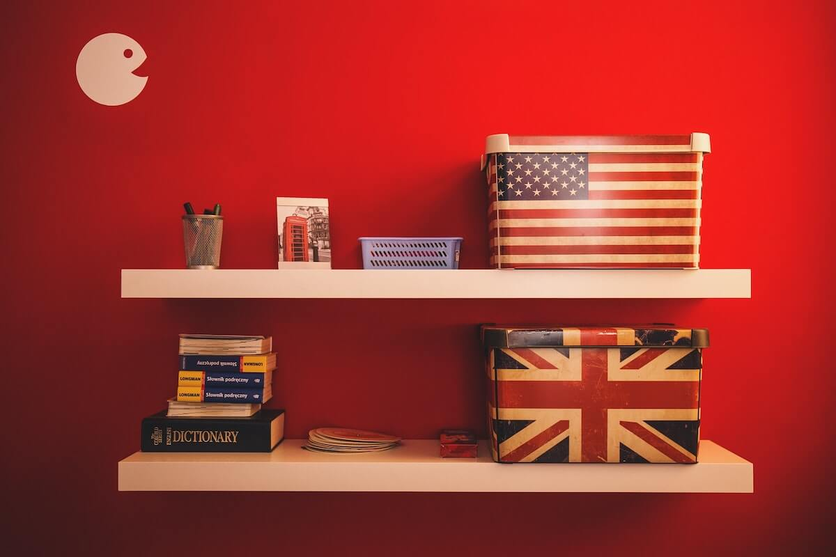 A pair of shelves with piles of books, including a dictionary, plus two boxes: one covered in the USA flag and one in the UK flag.