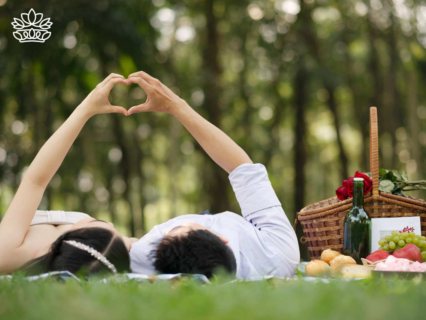 Couple forming a heart shape with their hands while lying on a picnic blanket in a lush park, with a picnic basket and romantic setup nearby, celebrating love in nature. Fabulous Flowers and Gifts. Delivered with Heart.