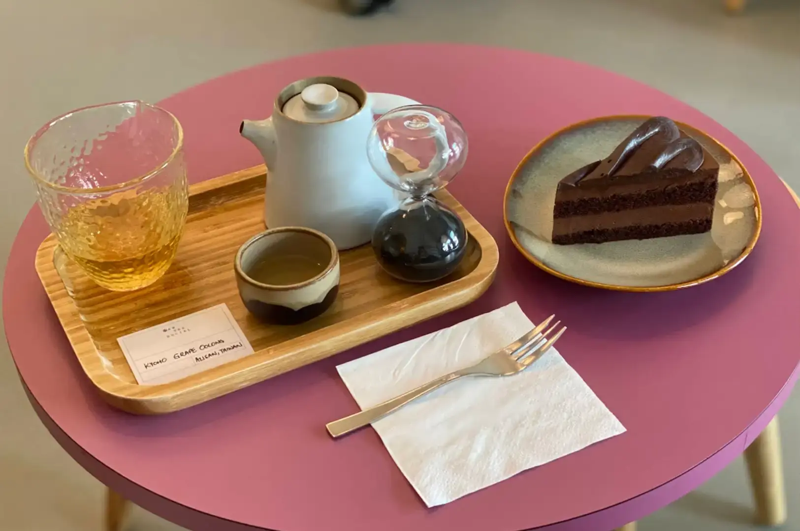 A high-angle view on a round, dusty-pink tabletop shows a slice of rich chocolate fudge cake on a ceramic plate set beside a wooden tea tray. The tray holds a white teapot, a small ceramic cup, a textured glass filled with iced golden tea, a small description card, and an hourglass tea timer with black sand. A silver fork rests neatly on a white paper napkin in the foreground.