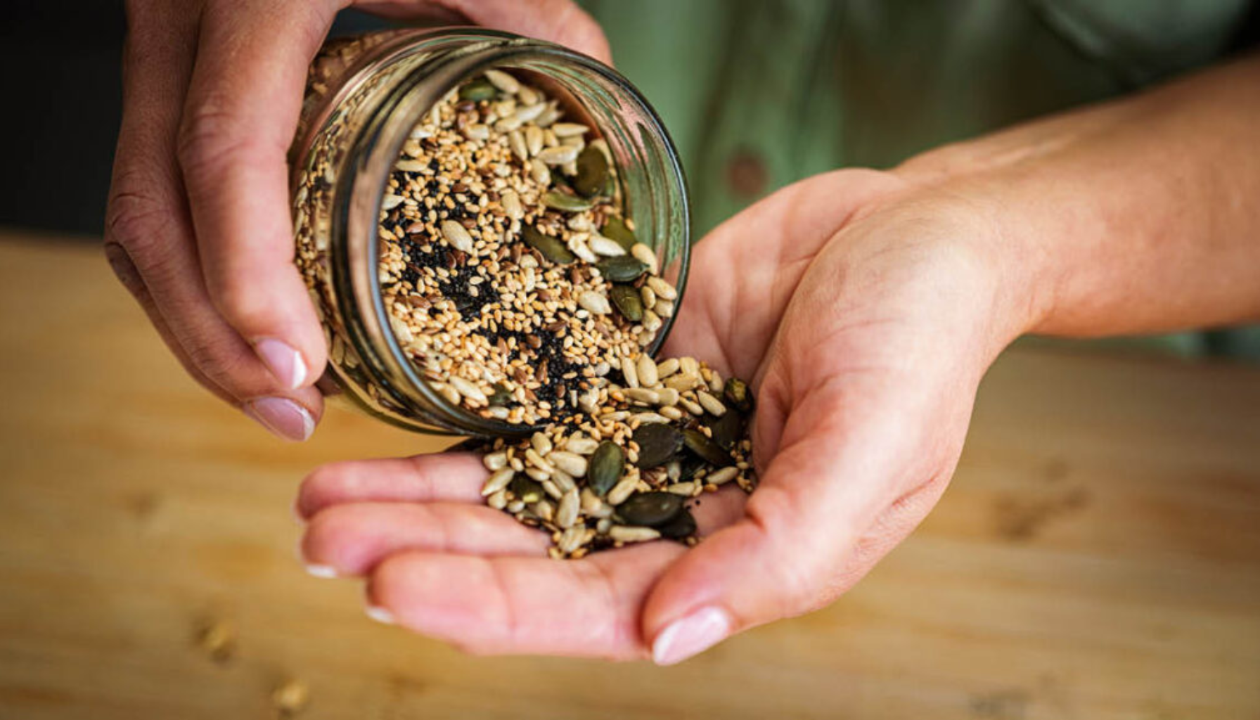 Hånd heller ut blandede frø som solsikkefrø, gresskarfrø og sesamfrø fra et glass over en annen hånd på kjøkkenbenk.