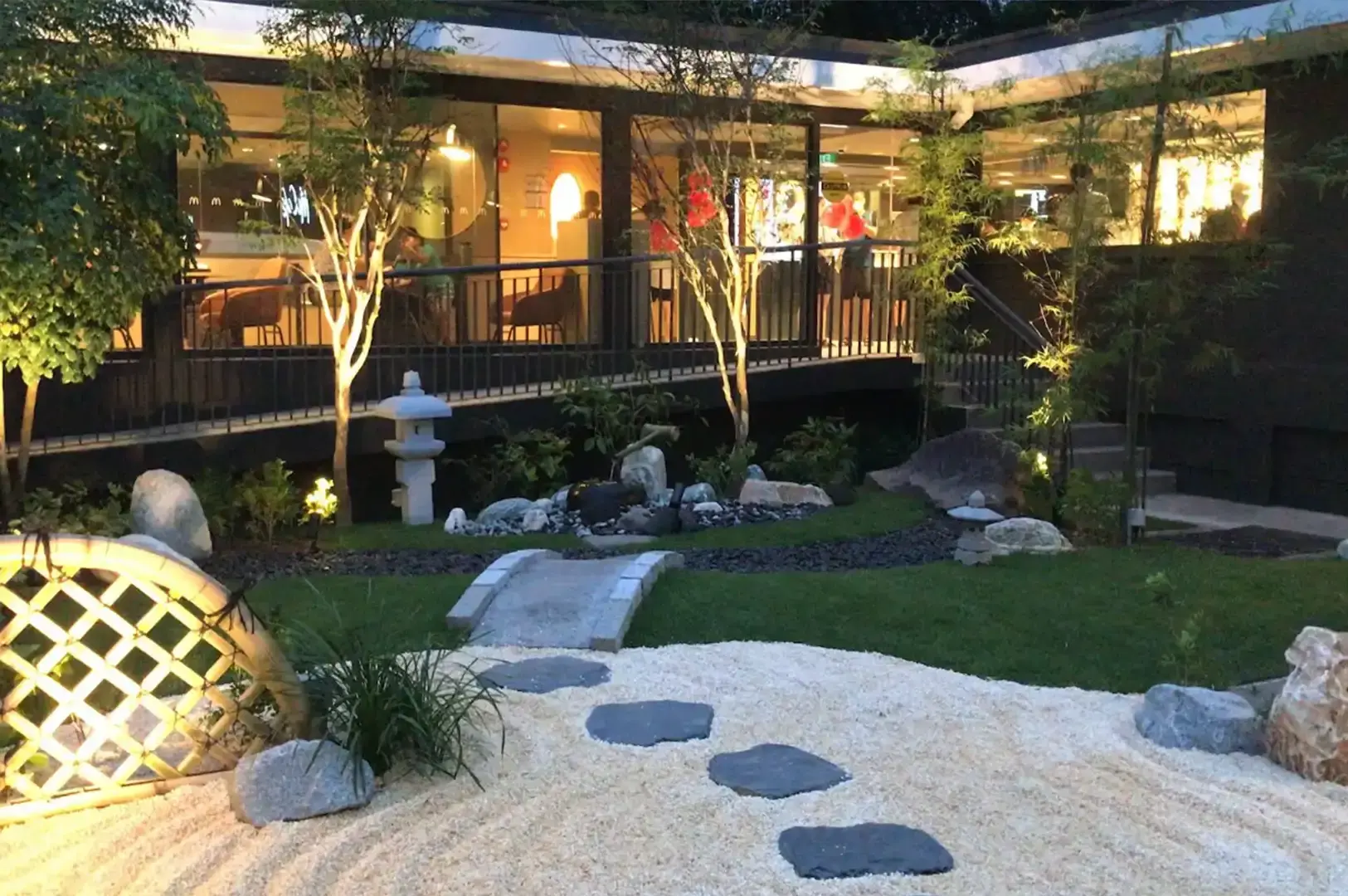 A warm evening shot of the zen garden area illuminated by soft uplighting, featuring a raked gravel bed with stone stepping-stones, a small stone arch bridge, decorative bamboo fences, and lit trees set in front of the glass-windowed, warm-lit restaurant interior.