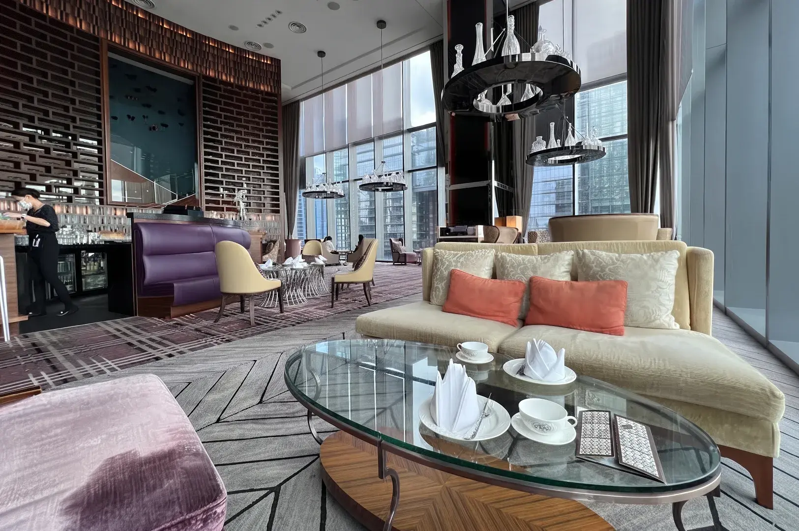 Elegant high-ceiling hotel lounge featuring cream and purple velvet seating, glass coffee tables set with teaware, and modern circular chandeliers against floor-to-ceiling windows.