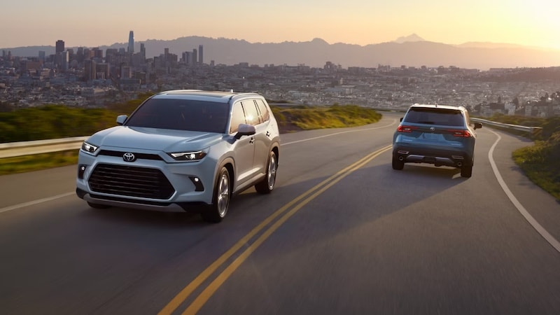 Two Toyota Highlander SUVs driving on a scenic road at sunset, highlighting their stylish design and performance, available at AutoFair Toyota of Tewksbury in Tewksbury, MA
