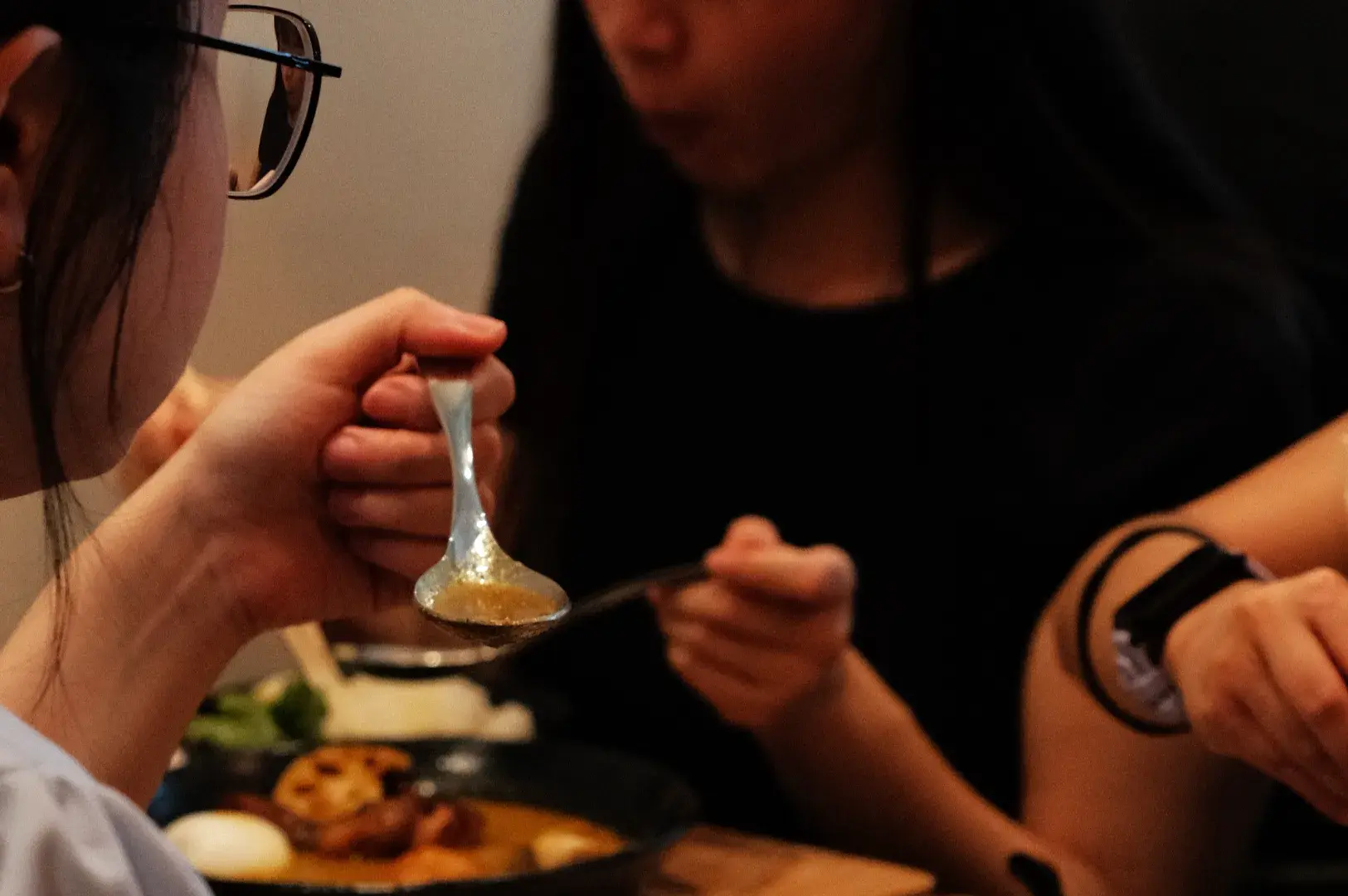 A close-up of a person holding a spoon, with others in the background enjoying soup curry.