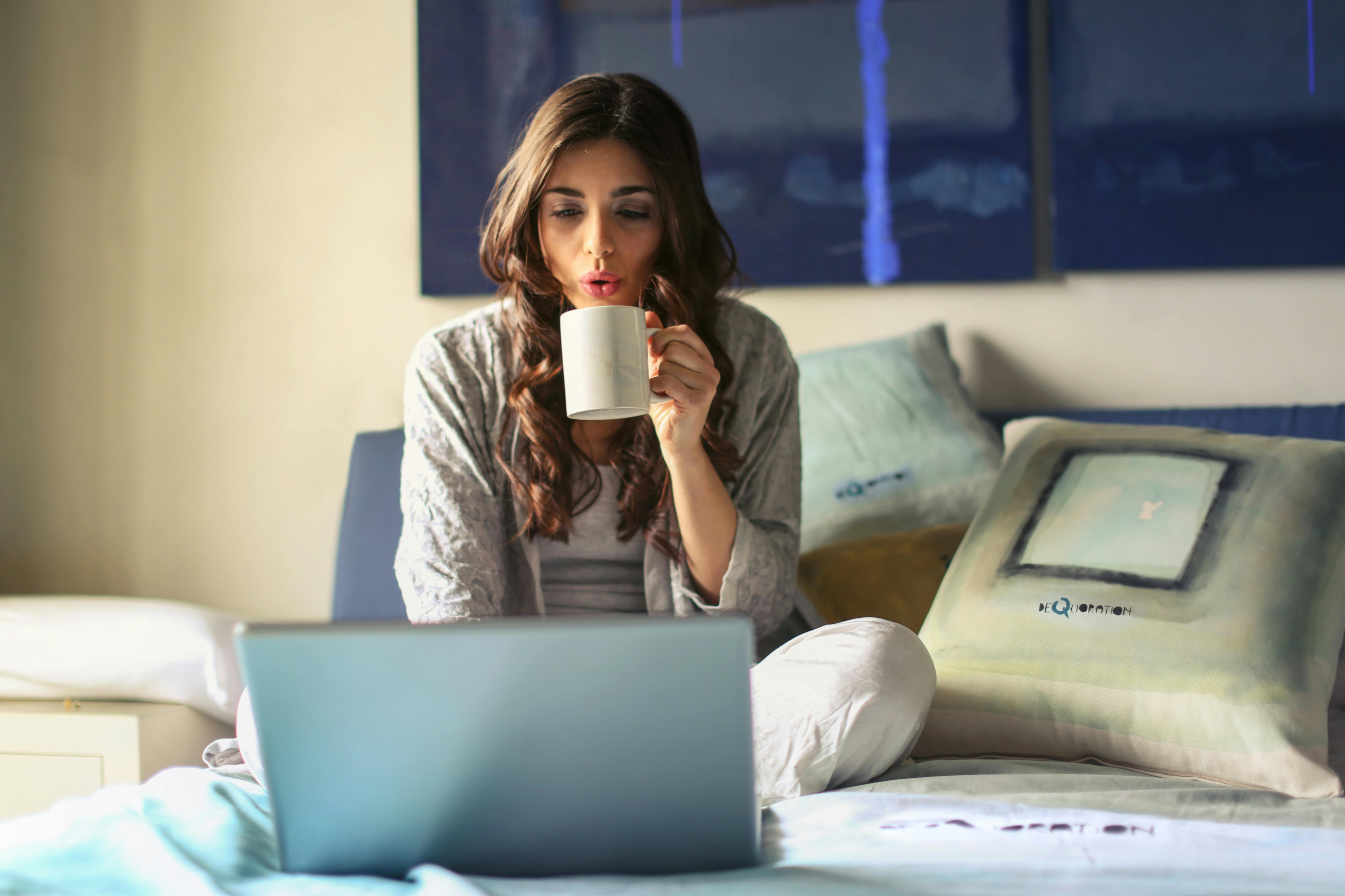 woman in grey sitting in front of a laptop in bed drinking coffee from a mug