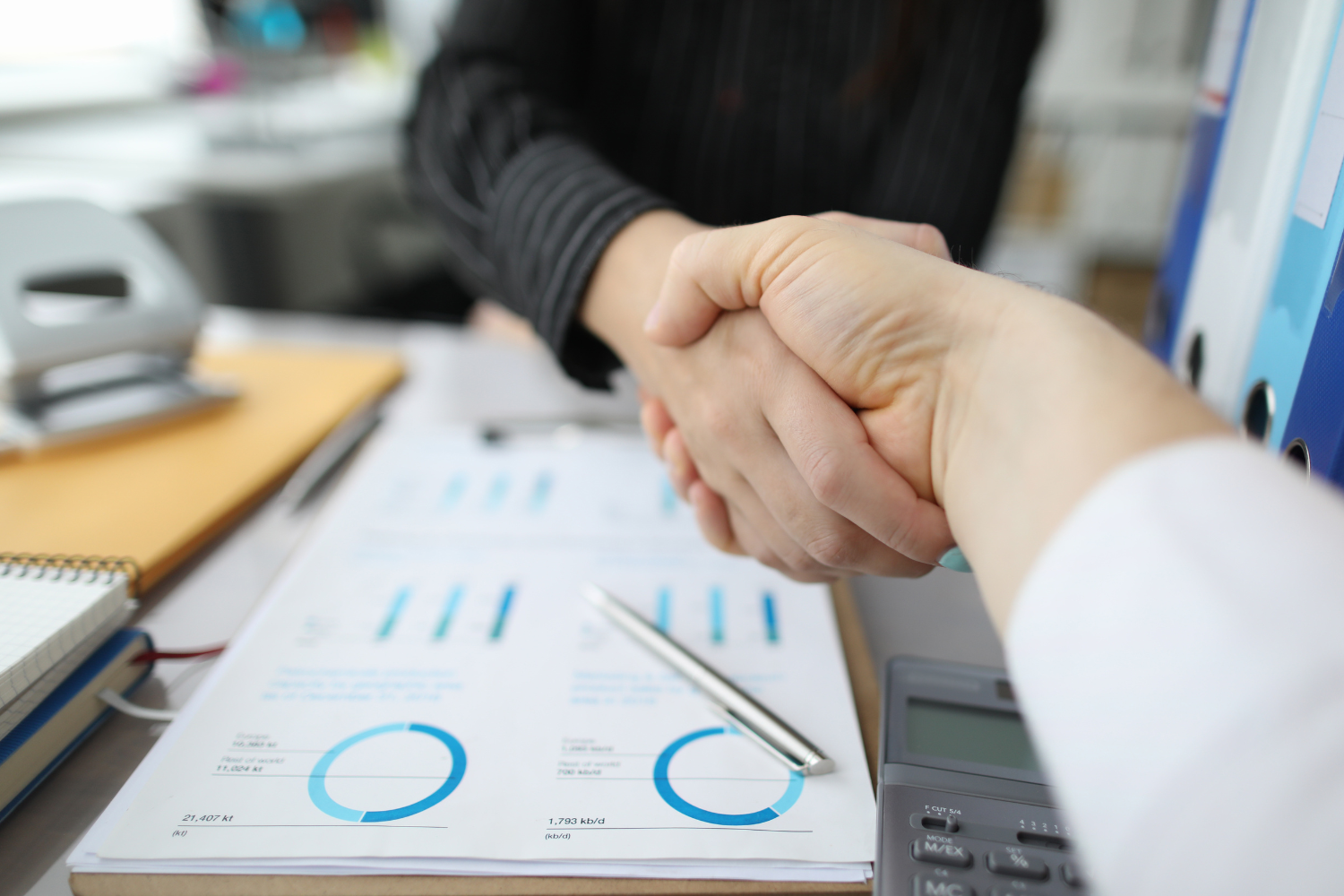 Two people shaking hands over a table with financial charts, a pen, and a calculator.