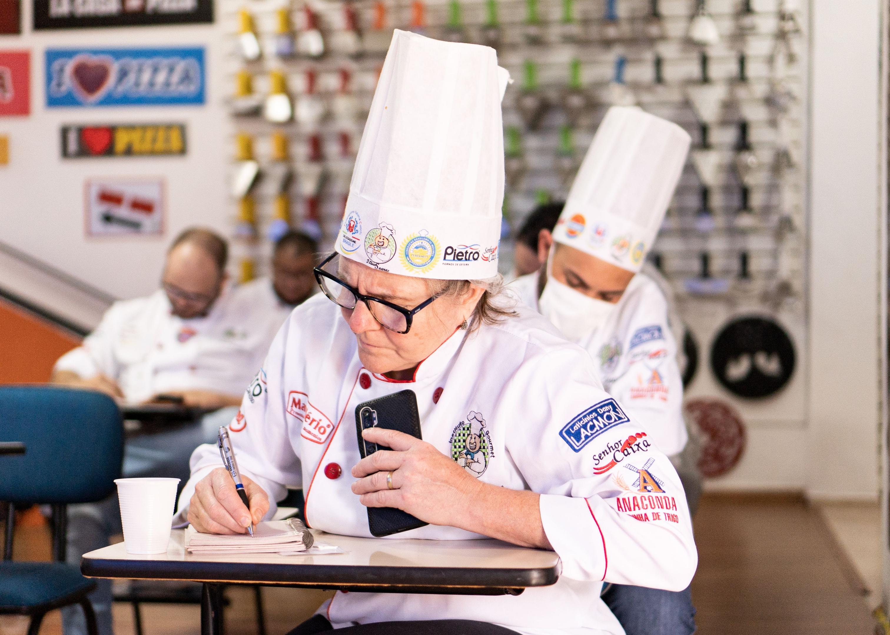 A restaurant chef in uniform holds a phone in one hand while writing an order on a notepad, illustrating how staff juggle incoming calls during service.