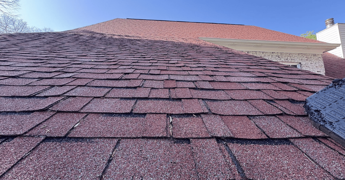 Red asphalt shingles with significant curling and lifting after prolonged exposure to direct sunlight and high summer temperatures.