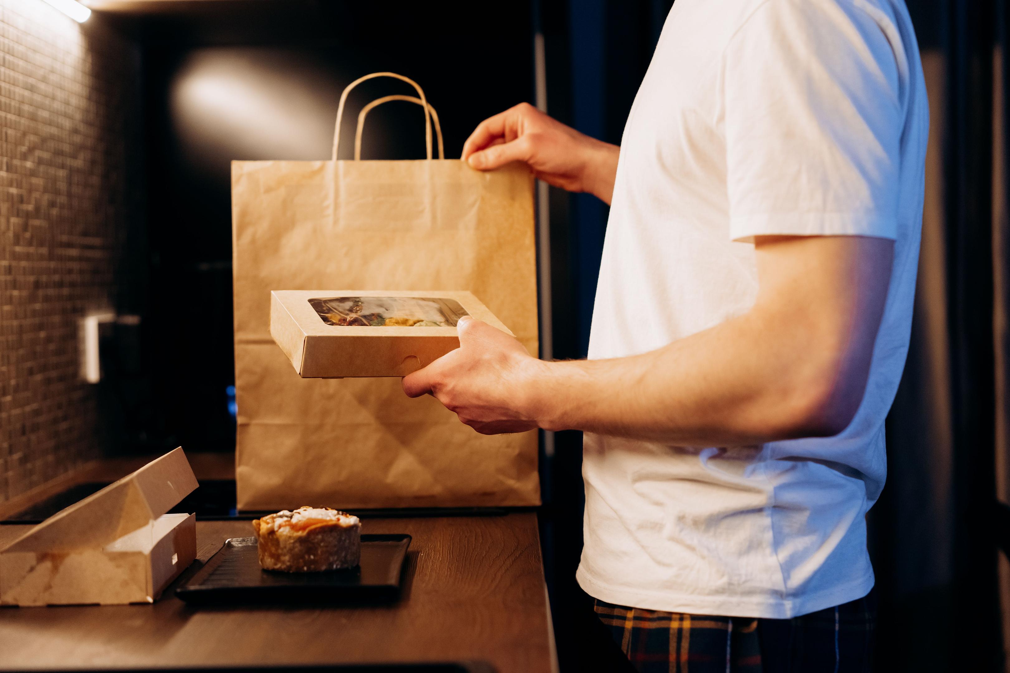 A person is holding a smartphone while standing near takeout bags on a restaurant counter, indicating the growing trend of online ordering in the restaurant industry. This scene reflects the importance of technology in enhancing guest satisfaction and attracting new customers for a successful restaurant business.
