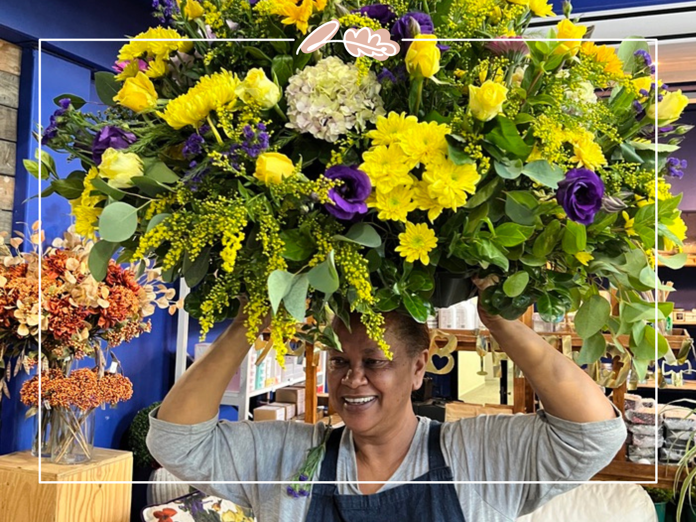 A florist balances a large, vibrant bouquet on her head in the store with pots for a function, capturing the joy and creativity in their work. Perfect for expressing love and care. Visit Fabulous Flowers and Gifts for more.