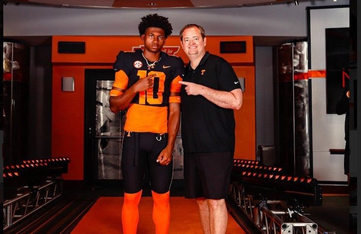 High school football recruit poses with a college coach in a locker room lit with team colors. The player wears a black and orange uniform with the number 10, while the coach smiles and points toward the recruit.