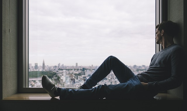 Man sitting on windowsill looking out at the big city. Sadness. When no career doesn't meet family expectations. 