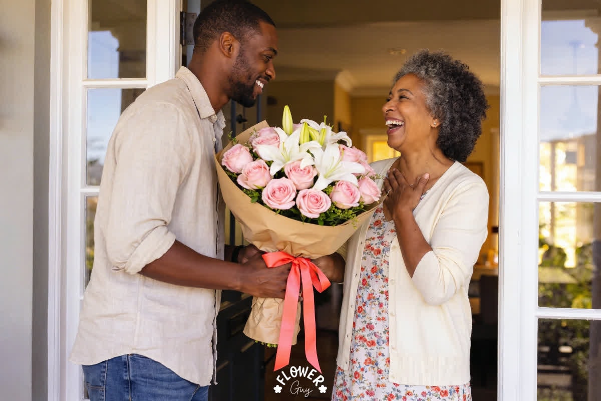 Young Black South African son surprising his mother with a bouquet of pink roses and white lilies wrapped in kraft paper with a coral ribbon at her front door, both laughing with joy