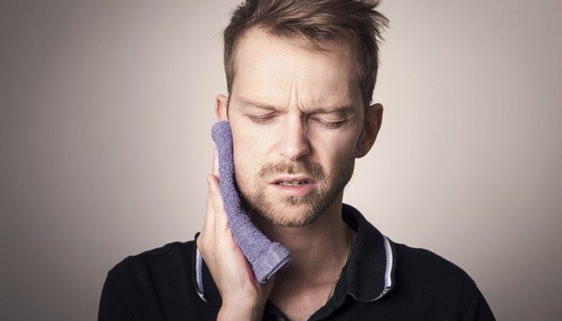 Mann med smertefullt ansiktsuttrykk holder en kald klut mot kinnet på grunn av tannpine eller kjevesmerter.