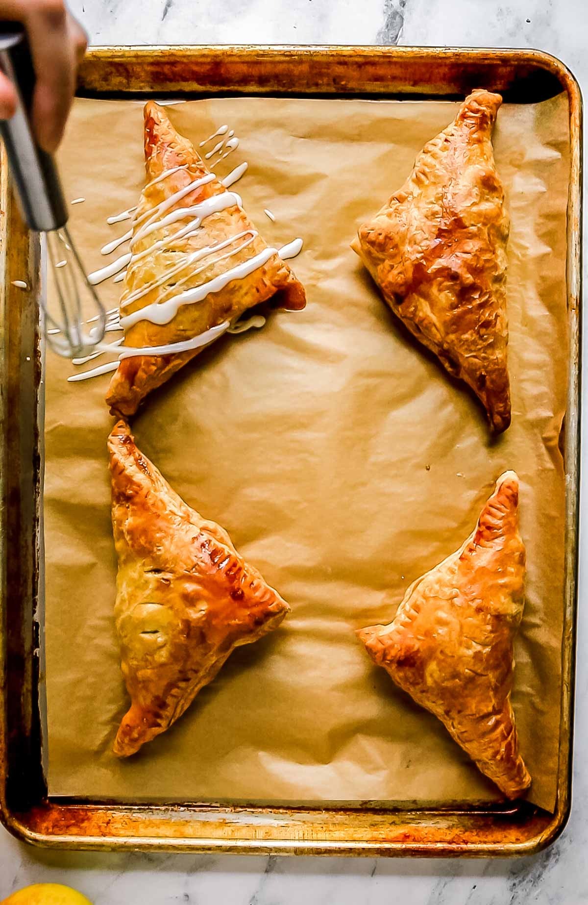 four puff pastry apple turnovers on baking sheet being drizzled with icing on parchment paper
