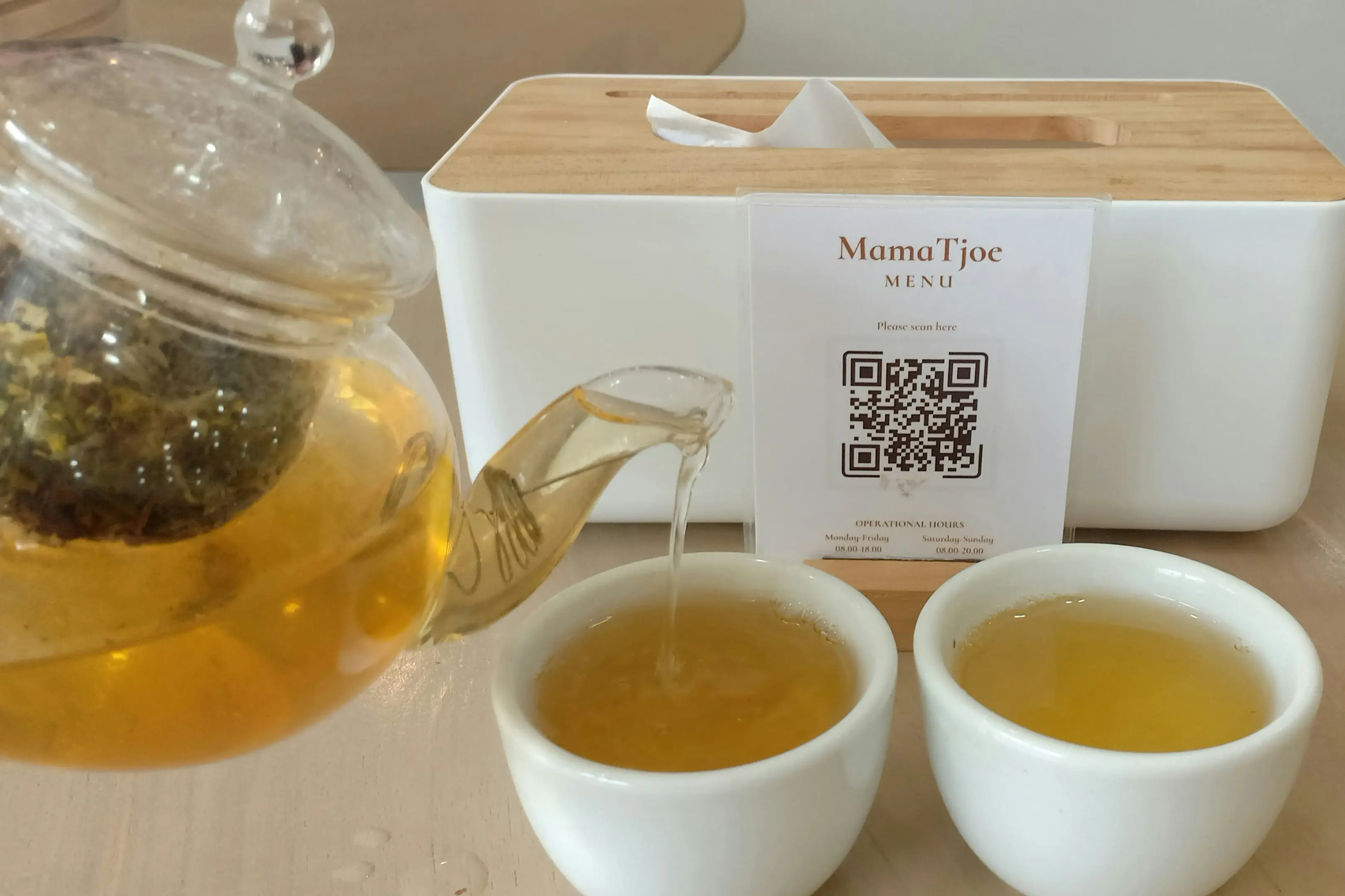 A clear glass teapot pours a golden-amber herbal tea into one of two small, white ceramic cups. In the background, a tissue box sits next to a small standing sign that displays a QR code for the "MamaTjoe MENU."