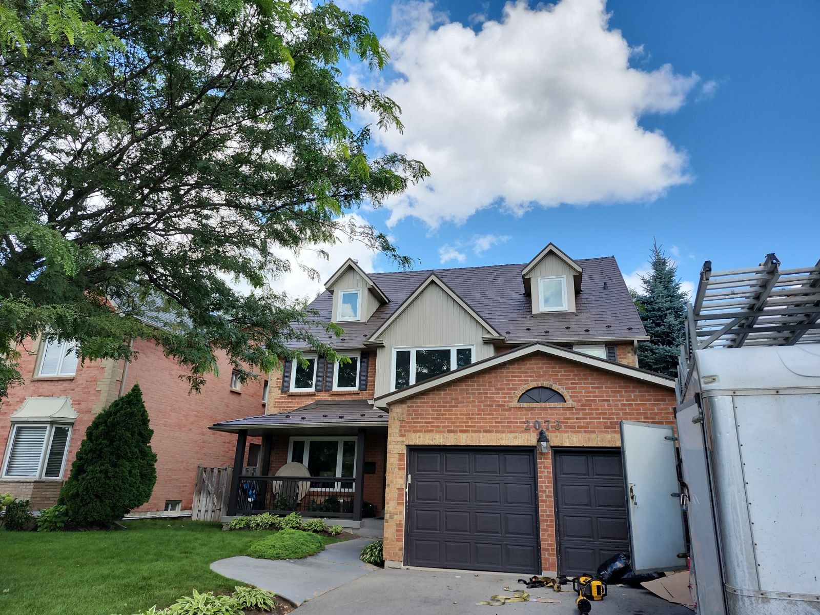 A brick house with a metal roof is surrounded by trees under a blue sky with fluffy clouds. A ladder leans against a truck trailer nearby. 