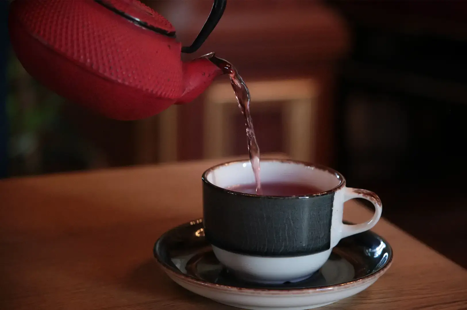 A close-up shot of a textured red cast-iron teapot pouring a thin stream of deep reddish-purple herbal tea into a dark ceramic teacup sitting on a matching saucer over a warm wooden tabletop.