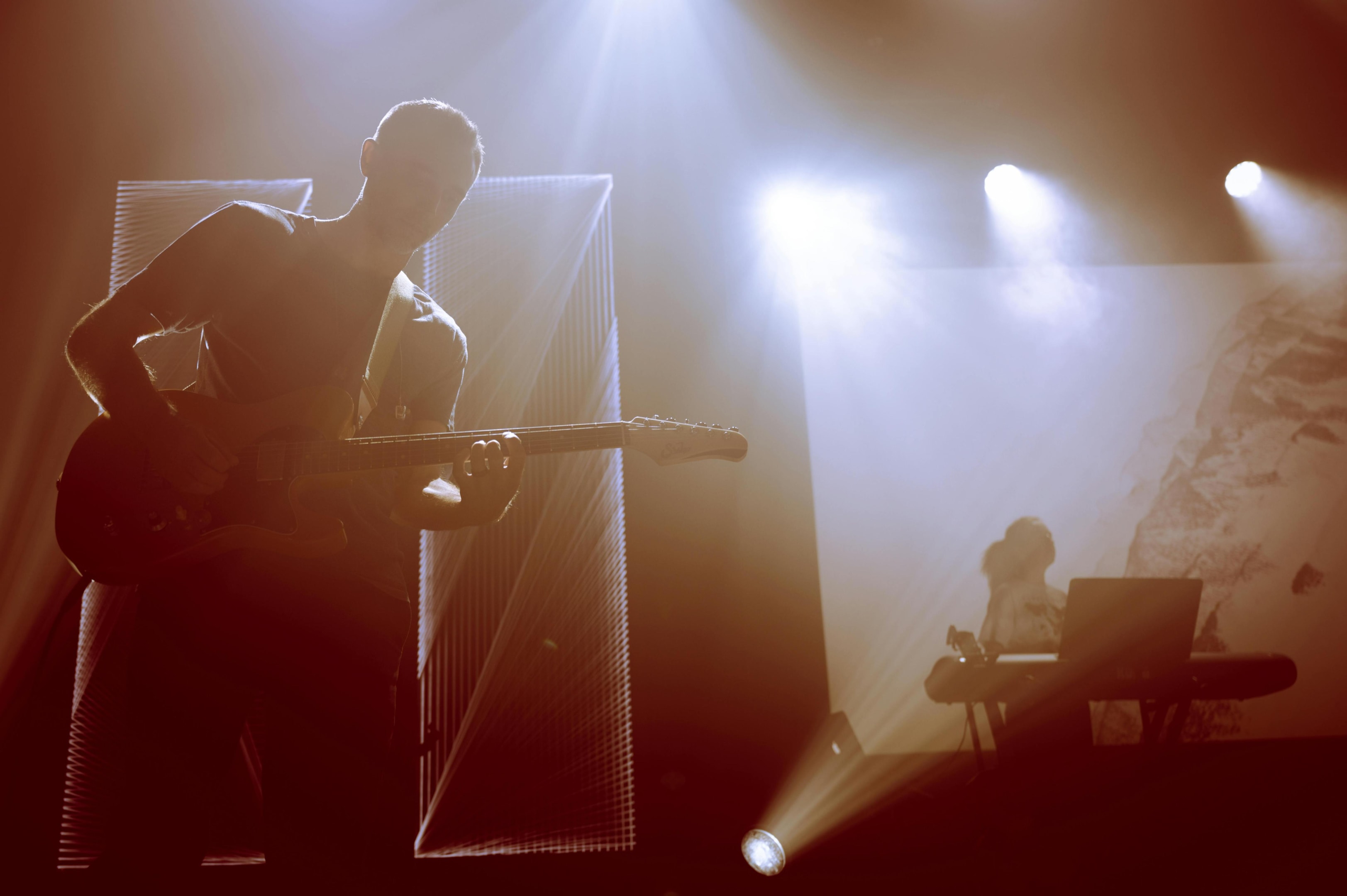 Backlit by dramatic stage lights, a male guitarist performs in silhouette, strumming an electric guitar with intense focus. Beams of light cut through the haze to reveal a keyboardist in the background, creating an atmospheric and energetic live concert setting.