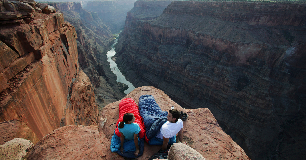 Two people in sleeping bags on the edge of a cliff overlooking a canyon - Adventure Wise Travel Gear
