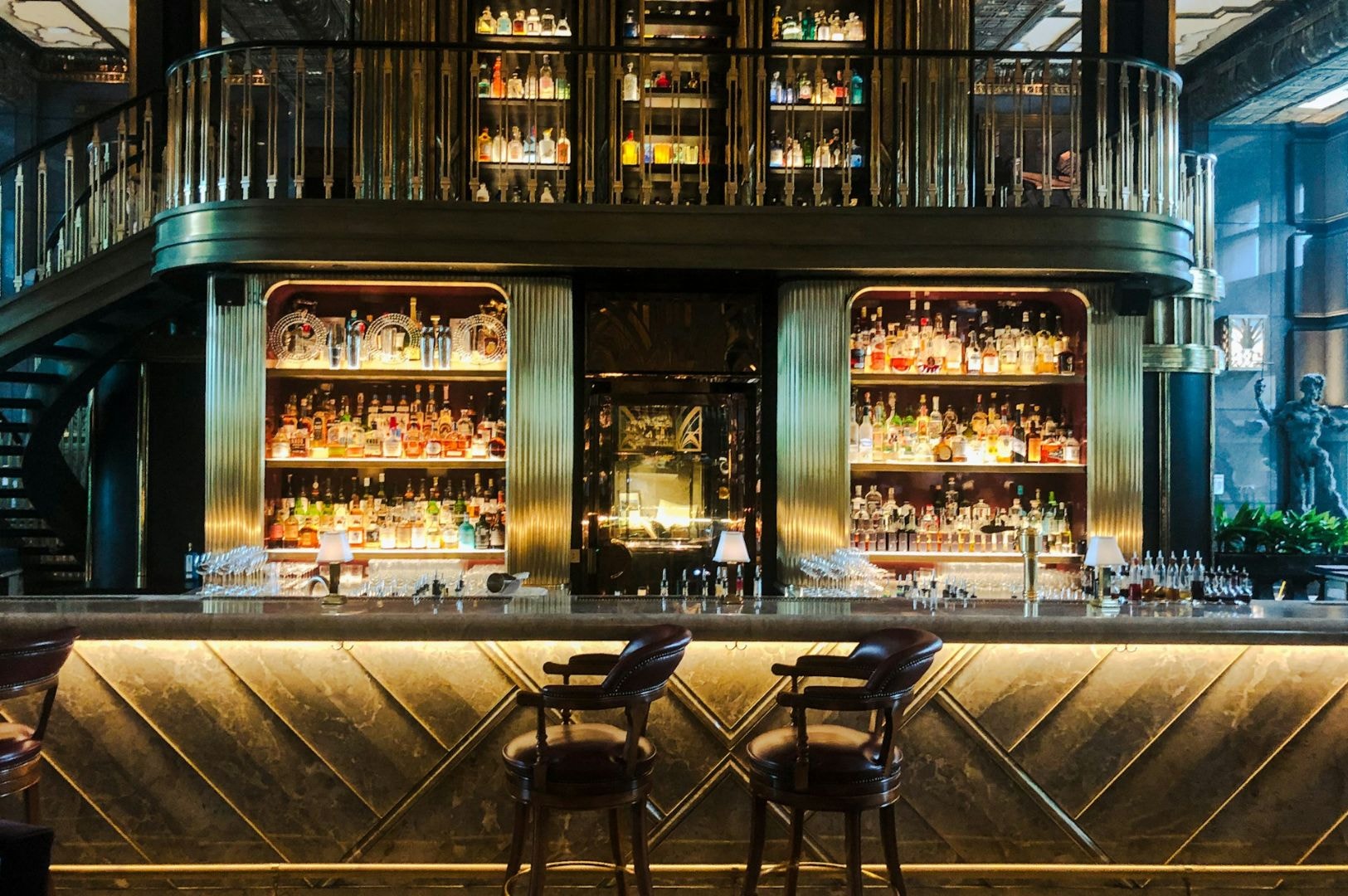 Elegant bar interior with a marble counter, leather stools, and warm lighting. Shelves behind filled with colorful bottles. A second-level gallery adds sophistication.