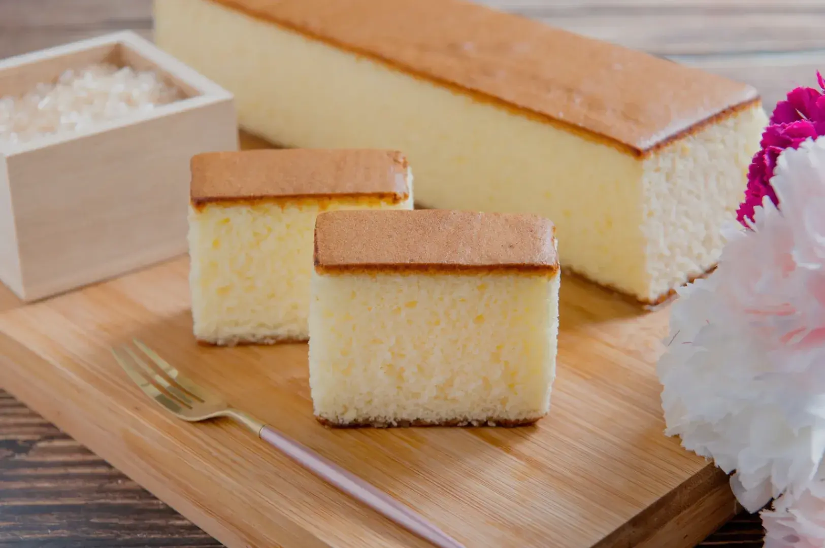 A wooden board with a sliced rectangular cake, two small pieces of cake, a box of sugar, and flowers.