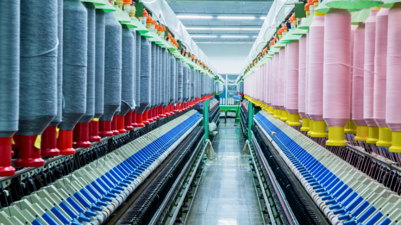 Rows of yarn spools in a textile factory, with gray spools on the left and pink spools on the right, illuminated by overhead lights.
