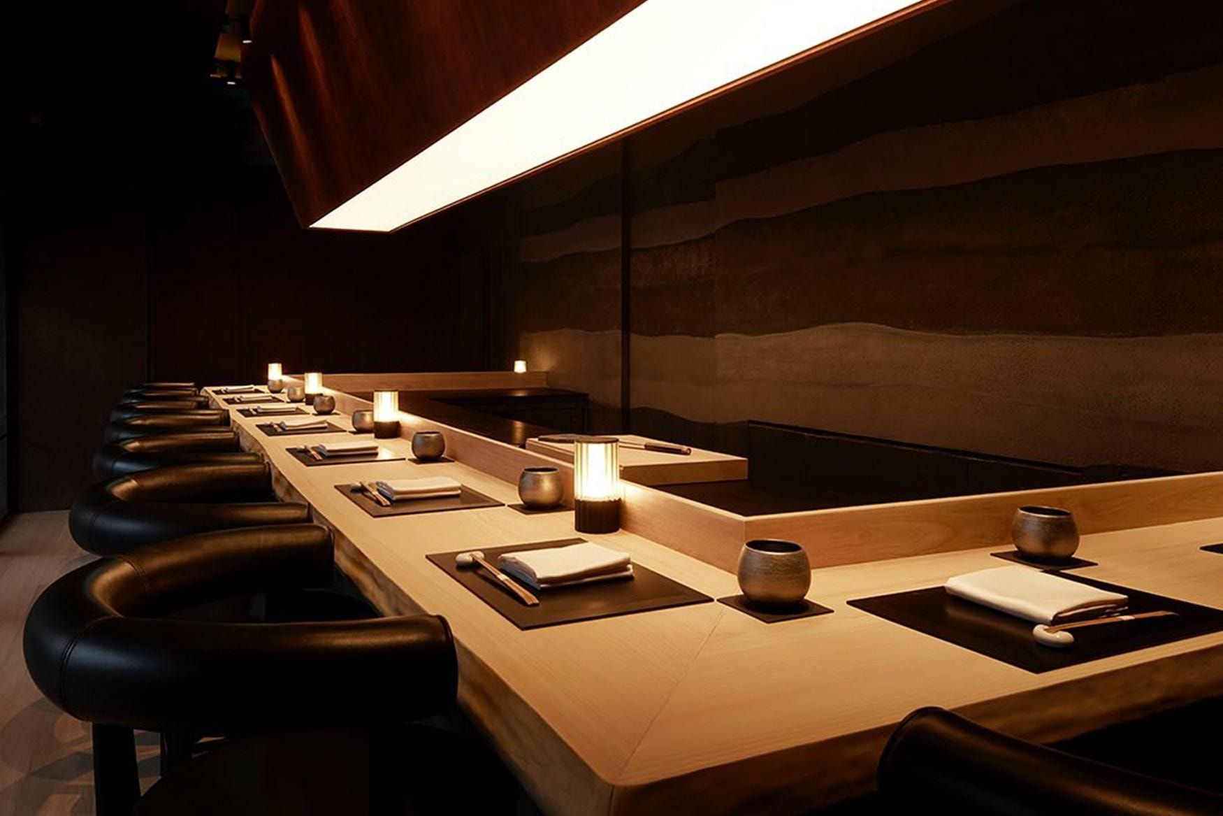  This image showcases a sophisticated, modern sushi counter featuring a sleek light wood surface, minimalist place settings, and curved black leather chairs. The atmosphere is intimate and moody, characterized by warm ambient lighting from small table lamps and a striking, angular overhead light fixture.