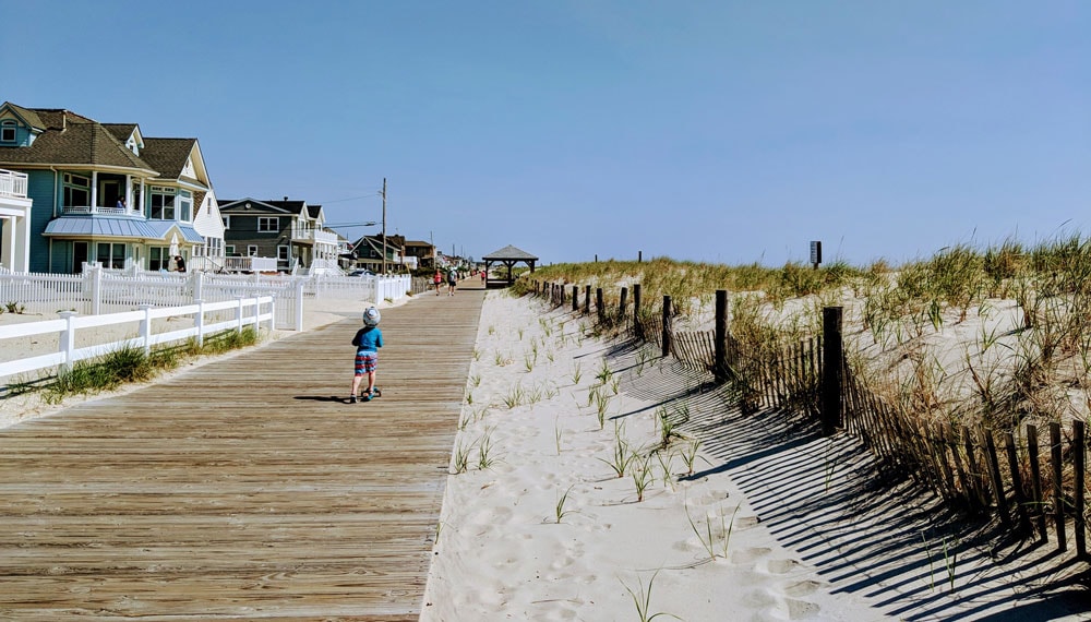 Child riding a scooter along the Lavallette boardwalk, with beach houses on one side and sand dunes with fencing on the other under a clear blue sky. Photo courtesy of Ocean County Business Development & Tourism.