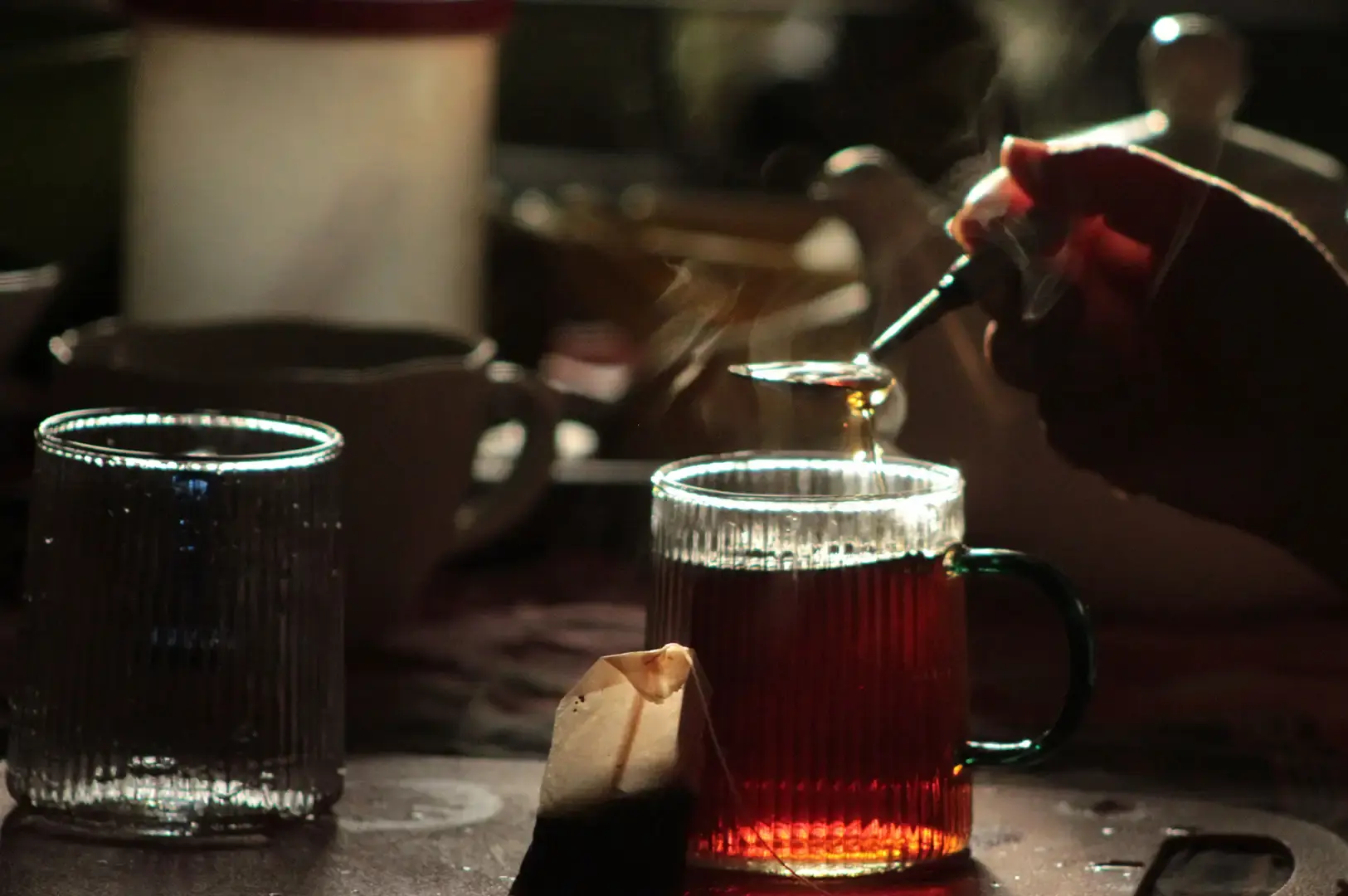 In a dimly lit setting, steam rises from a clear ribbed glass mug as dark liquid is carefully poured in, with a paper teabag resting on the wooden counter beside it.