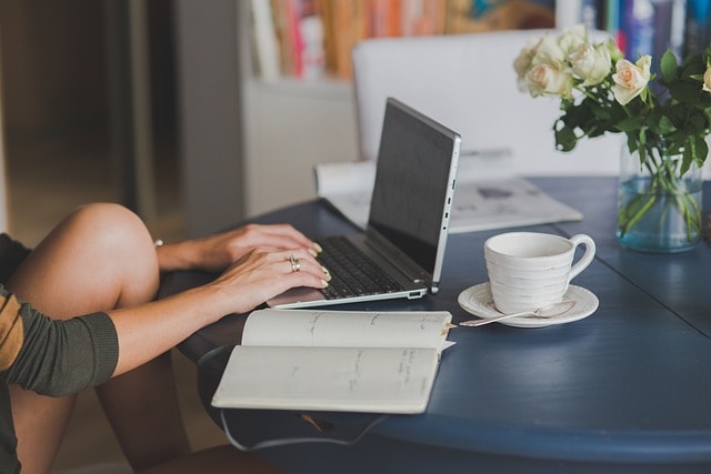 A woman typing on her notebook with an open journal next to her and a cup, representing the life of a writer.