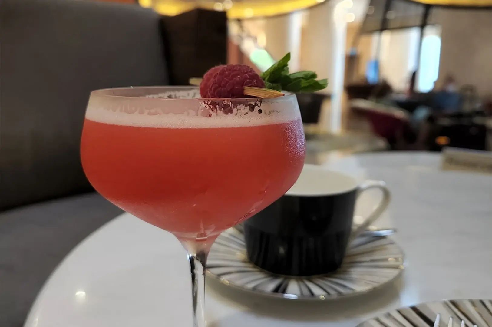 A vibrant, pink-hued cocktail in a stemmed coupe glass is garnished with a single fresh raspberry and a sprig of mint, serving as a bright focal point on the table. Behind the cocktail, a sophisticated black and white striped teacup and saucer are partially visible, set against the soft-focus, luxurious interior of the hotel atrium, capturing a refined and indulgent afternoon experience.