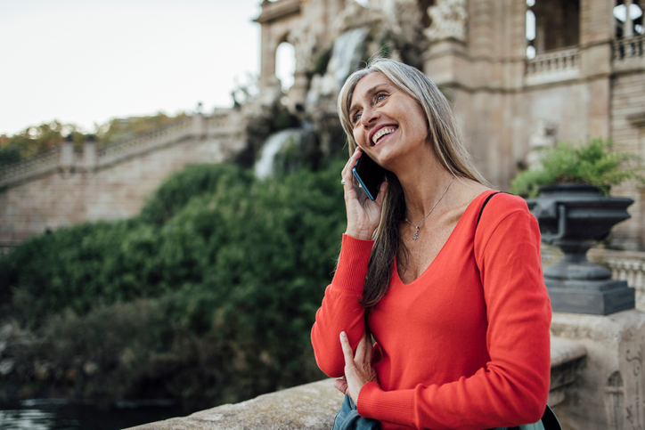 Happy women with long grayish hair chatting on her cell in front an ancient building. 
