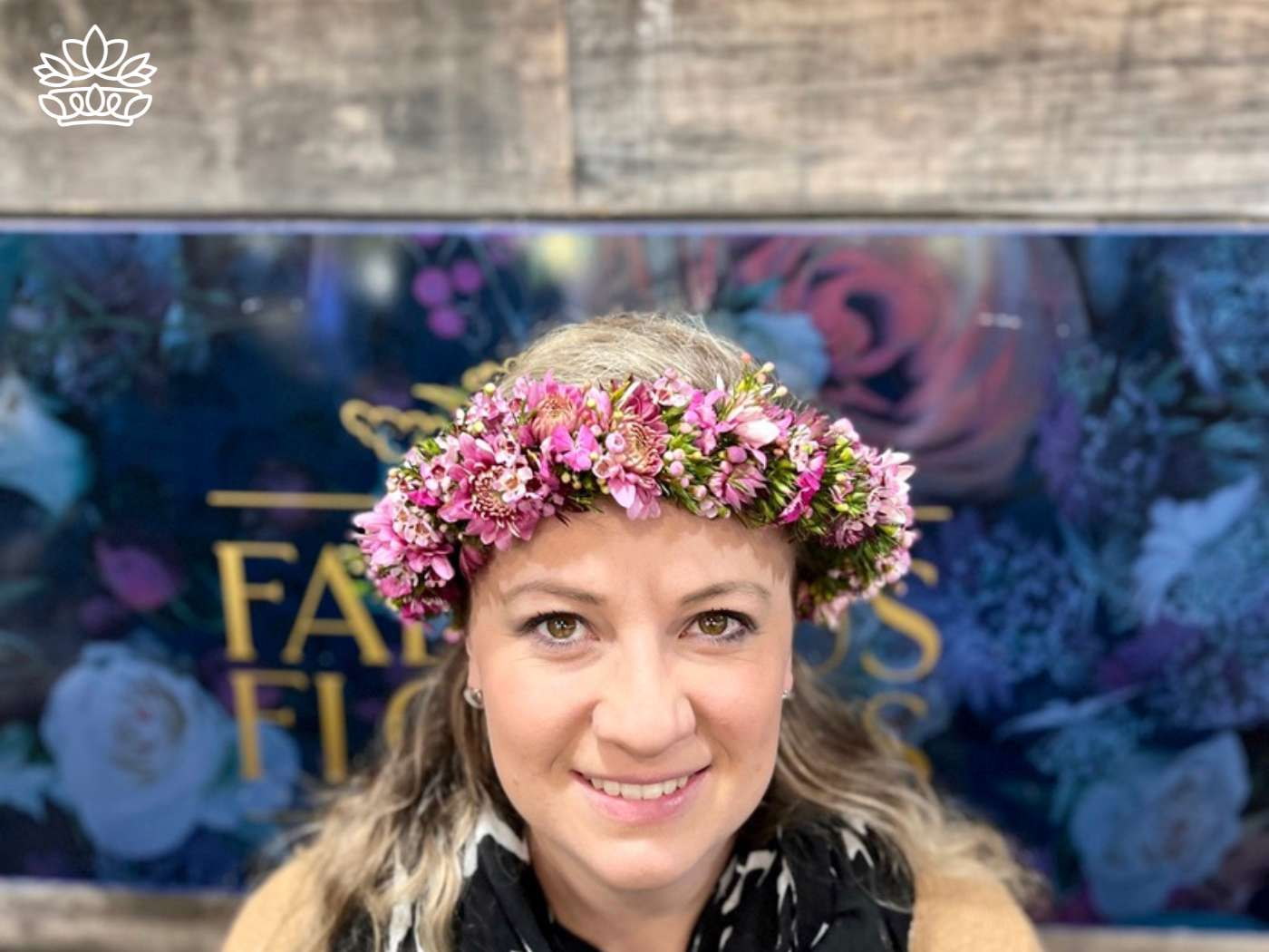 A smiling woman adorned with a vibrant pink floral crown against a backdrop with 'Fabulous Flowers' text, representing the festive and handcrafted selection from Fabulous Flowers and Gifts.
